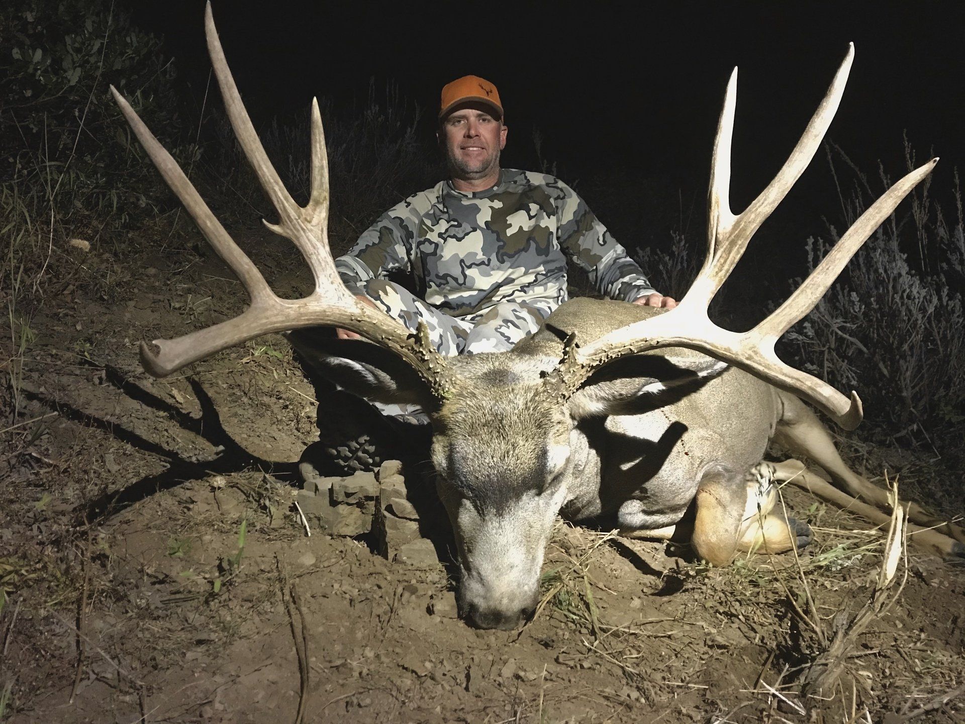 Hunter with a large buck deer, posed outdoors at night. Man wears camo and an orange cap.