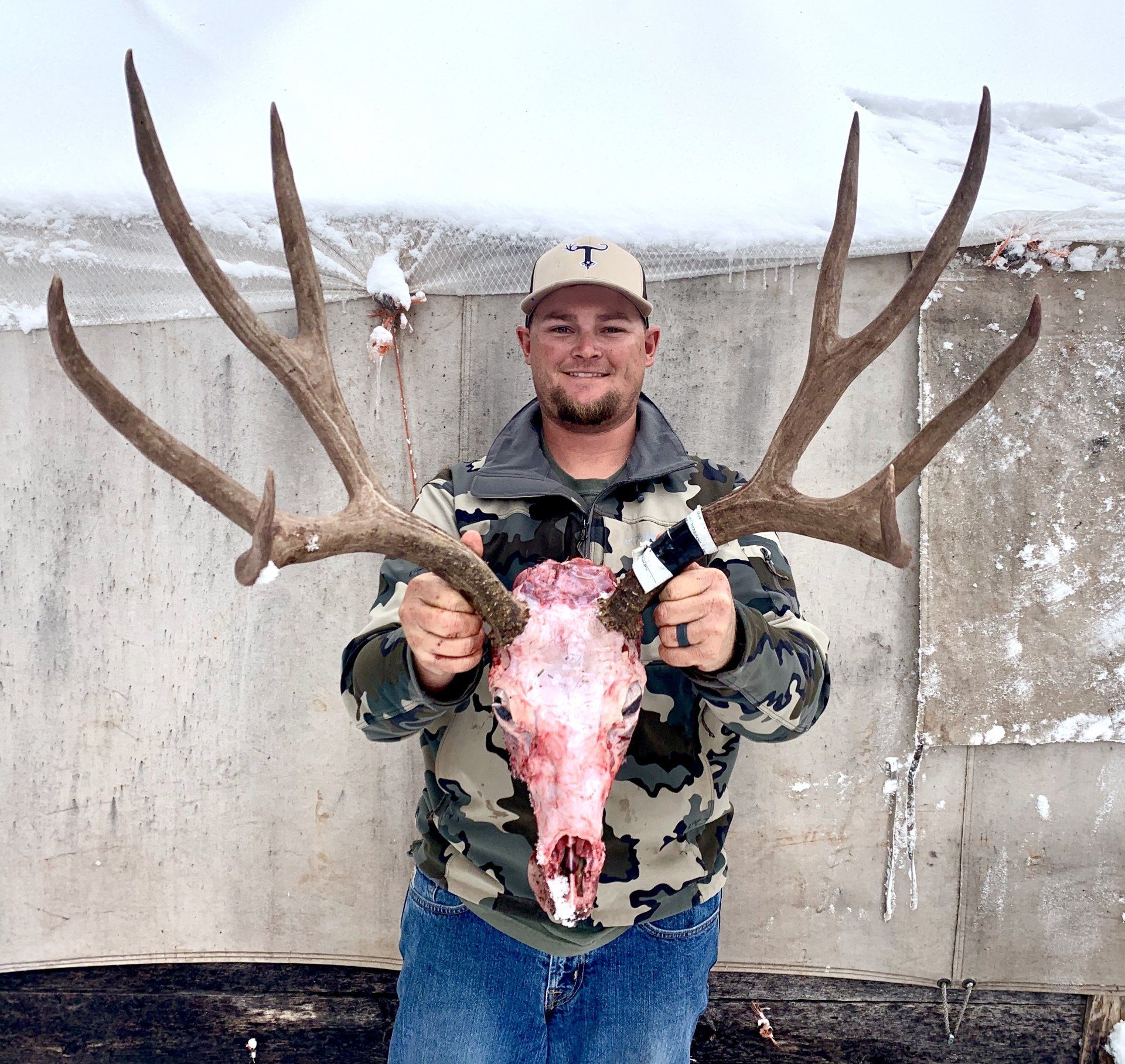 Man holding elk skull and antlers, smiling in front of a concrete wall.
