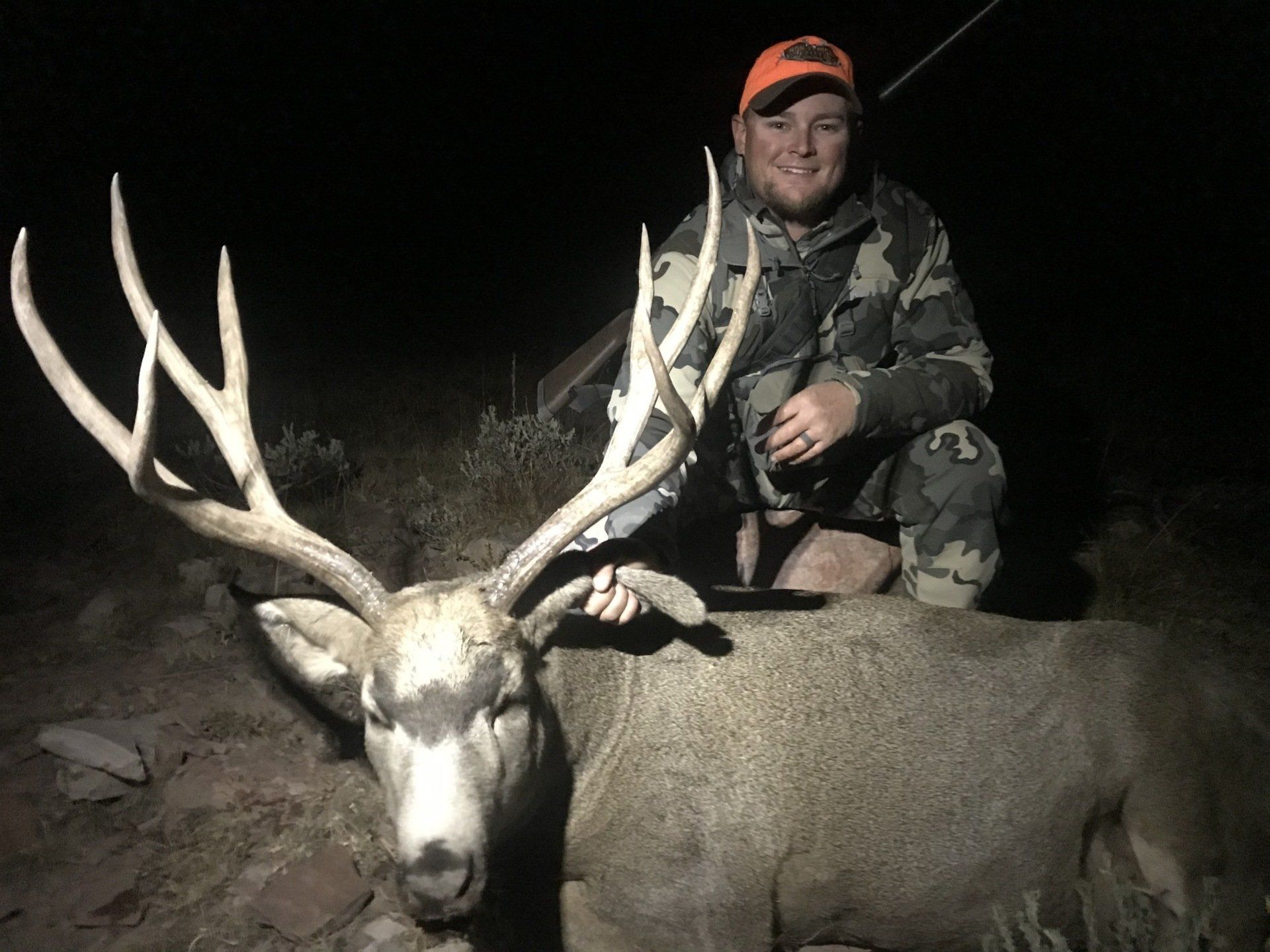 Hunter kneels next to a large buck with impressive antlers at night; wears camouflage, orange hat.