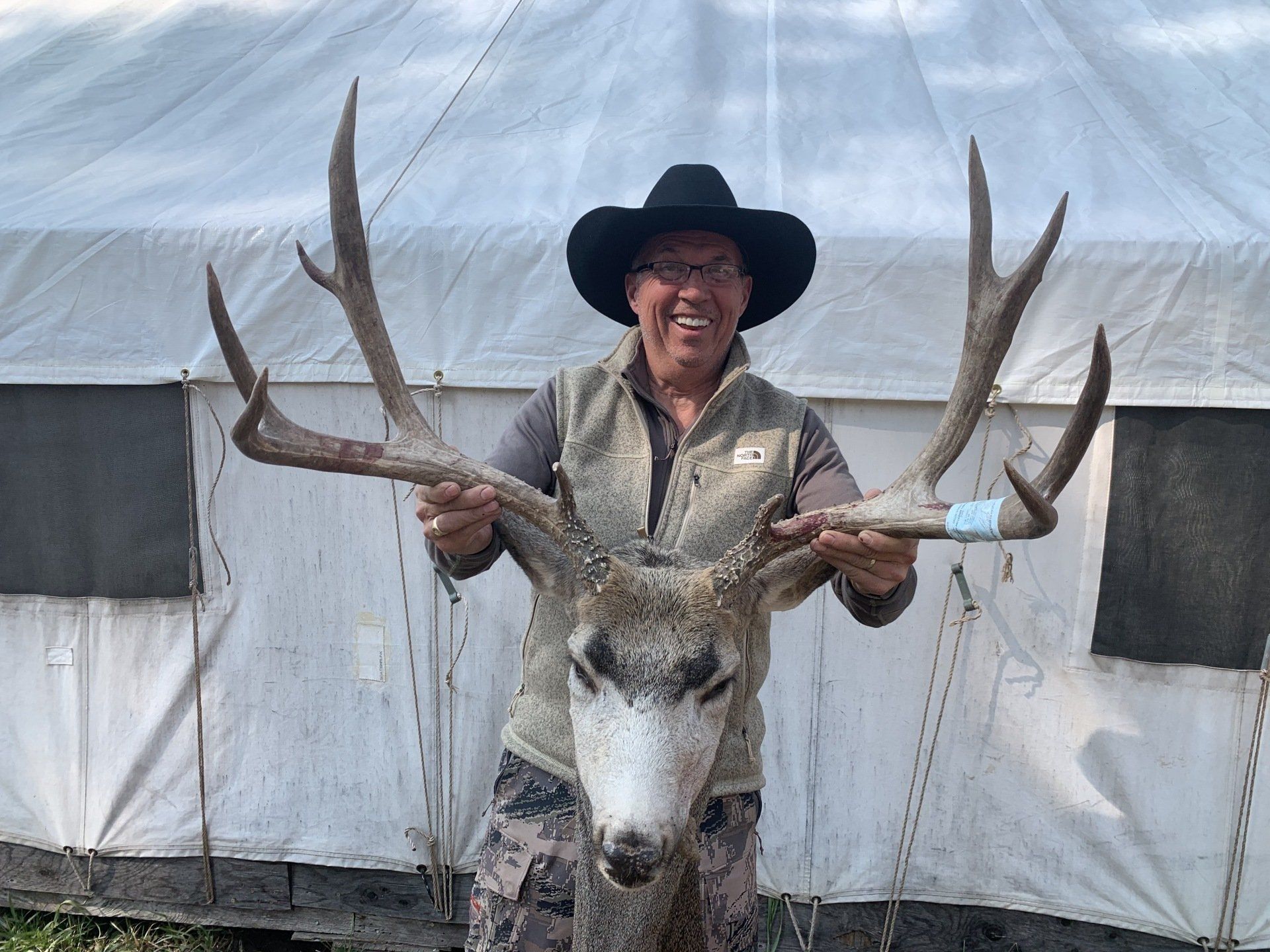 Person wearing a cowboy hat smiling, holding large deer antlers in front of a tent.