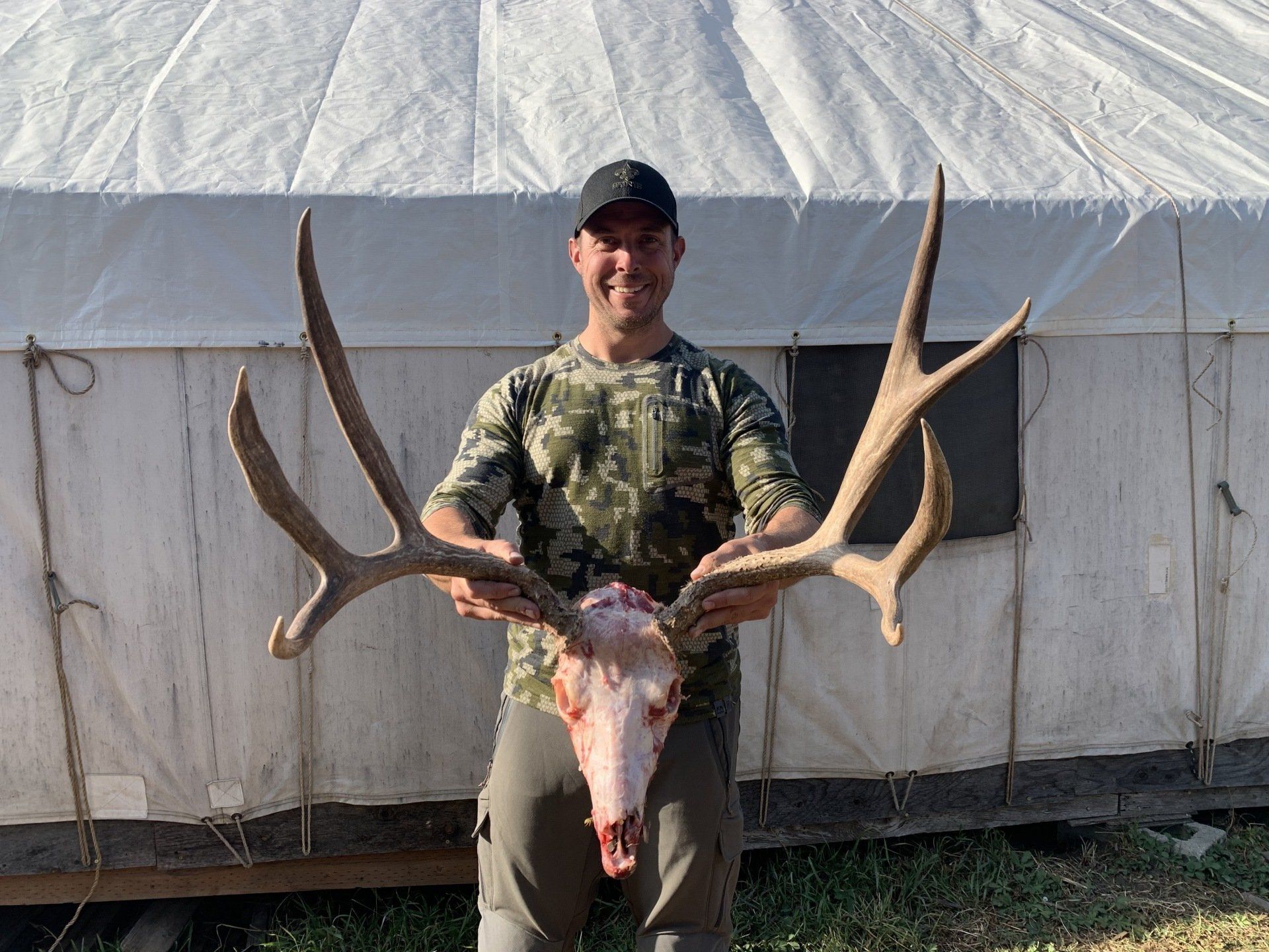 Man holding elk skull and antlers in front of a white tent. Smiling. Outdoors.
