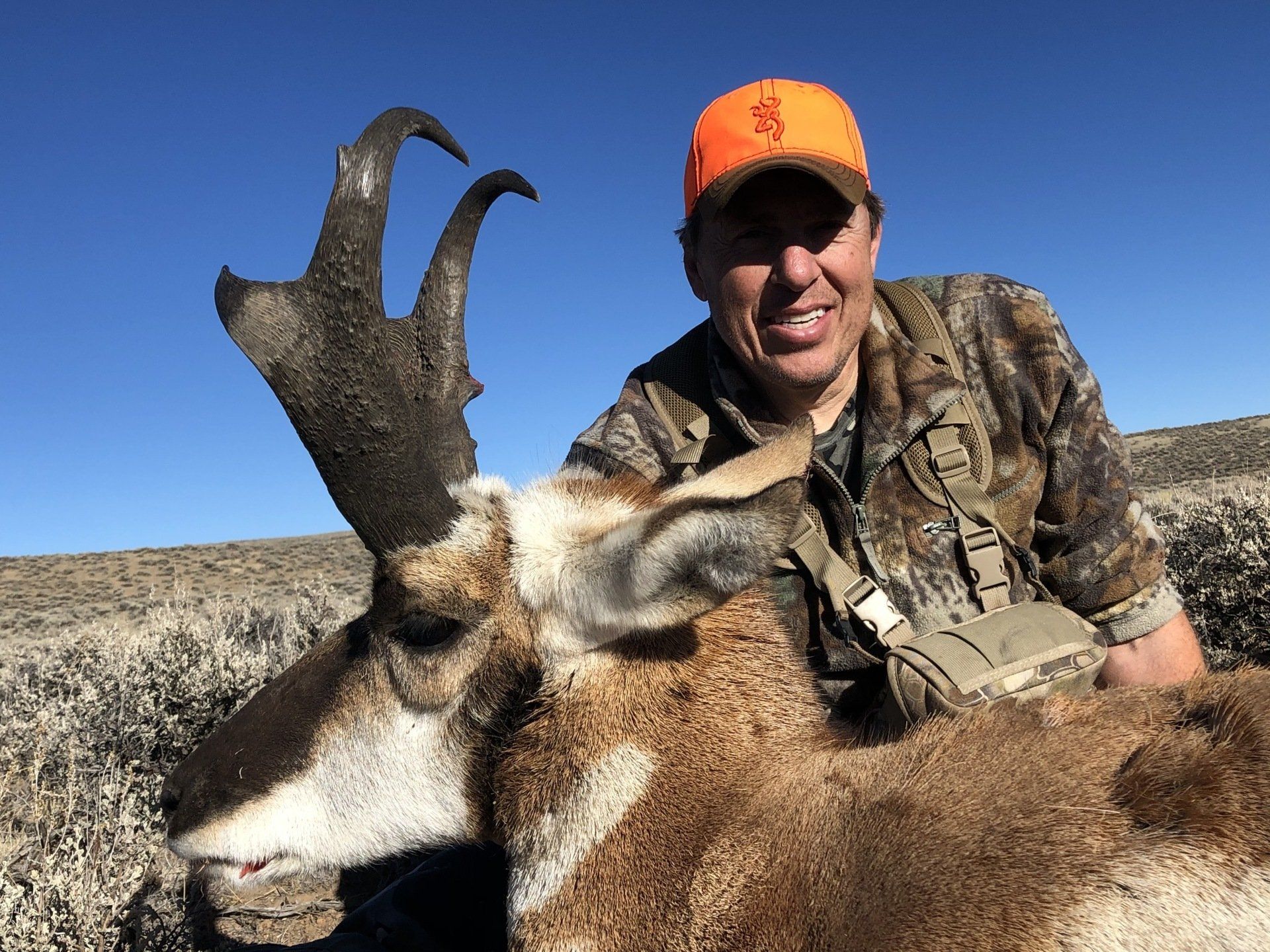 Man in camo, orange hat, smiling, with a trophy pronghorn antelope on the ground; blue sky, arid landscape.