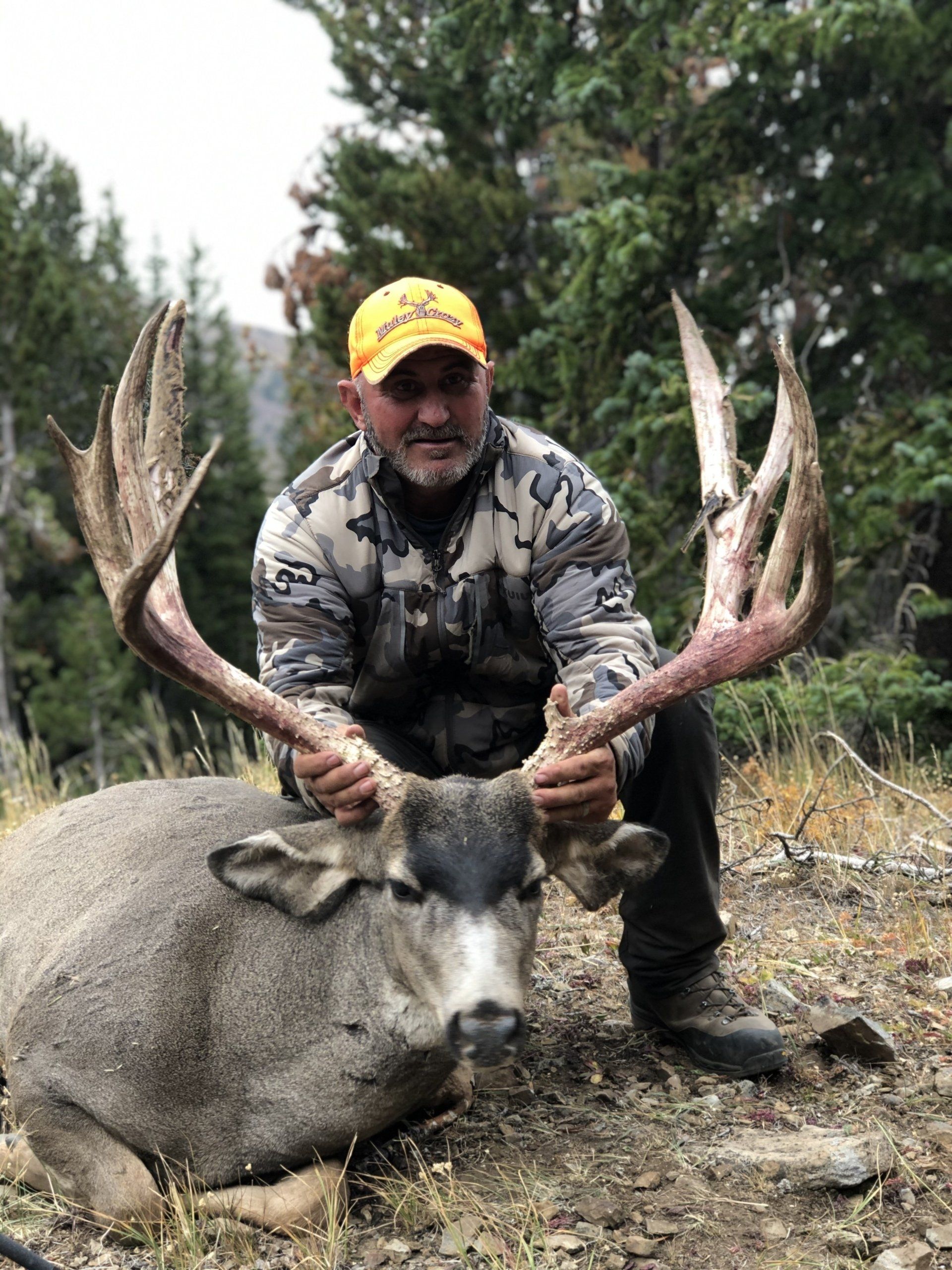 Man holding large deer antlers, posing next to harvested buck in forested area.