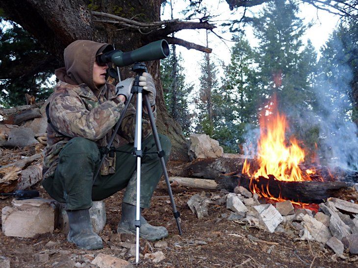 Person wearing camouflage uses a spotting scope on a tripod near a campfire in a forest.