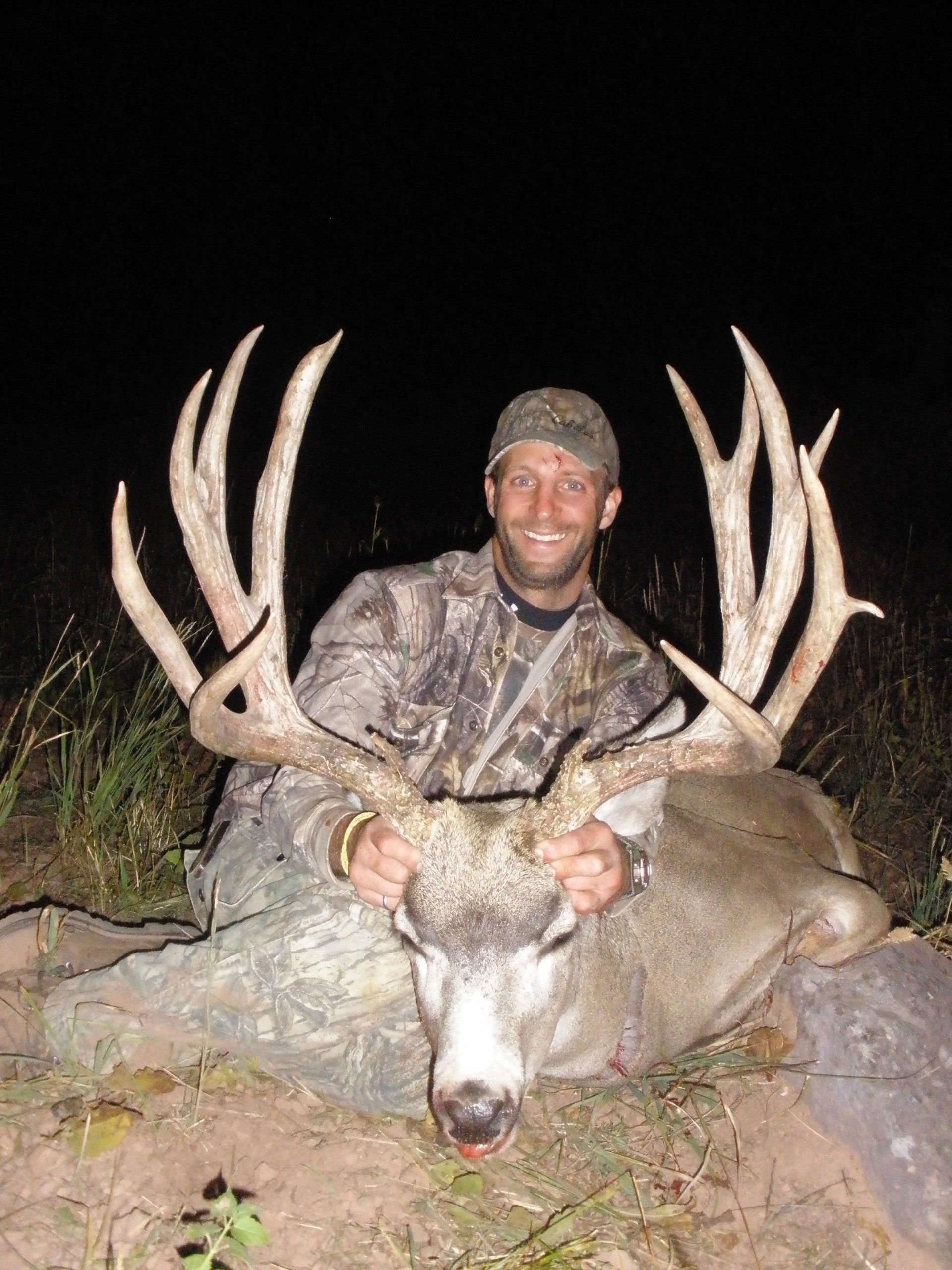 Man in camo holds large deer antlers; nighttime, smiling.