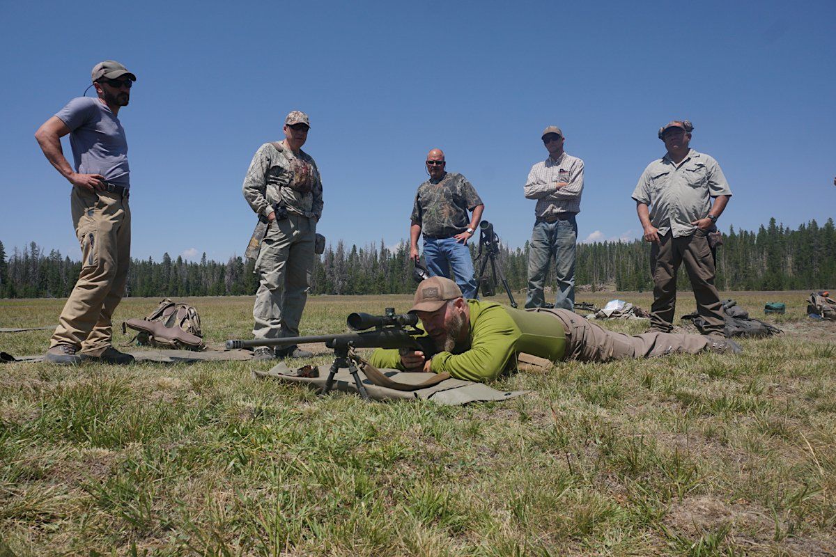 A person prone on the ground aiming a rifle, with five people standing behind on a grassy field on a sunny day.