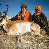 Two people pose with a pronghorn antelope on a grassy plain. Both wear hunting vests and hats.