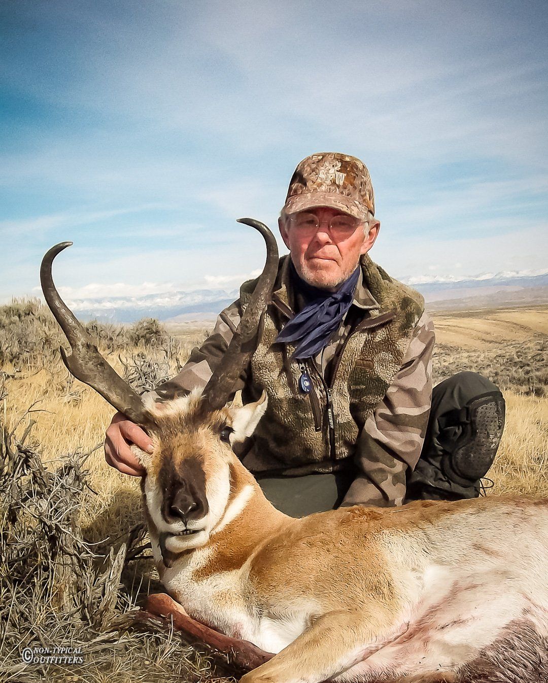Hunter kneels beside a pronghorn, holding its large horns. Brown and tan camouflage clothing. Outdoors, in a field.