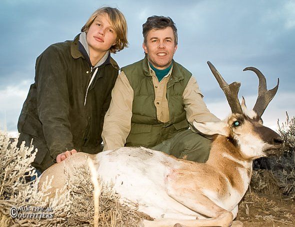 Two people pose with an antelope. One is petting it. Brown, tan, and green clothing in an outdoor setting.