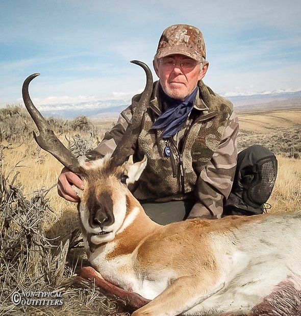 Hunter kneels beside an antelope in a field, holding its antlers. He wears camo clothing, with mountains in the background.