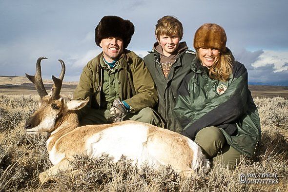 Family posing with a pronghorn antelope in a field; they wear hunting attire.