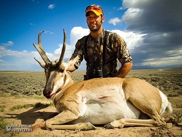 Hunter posing with a harvested pronghorn antelope in a desert setting; blue sky and clouds.