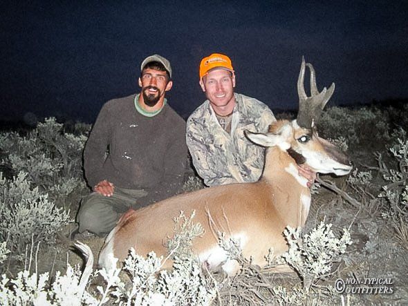 Two men pose with a pronghorn antelope in a field, one kneeling. One man wears camo, the other a green shirt.
