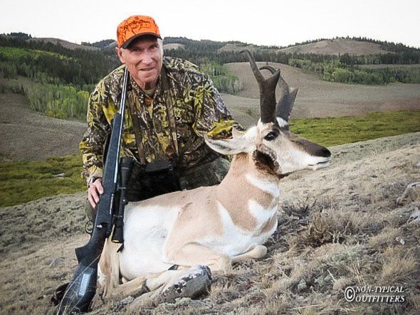 Hunter in camouflage with orange hat, and  Wyoming trophy buck pronghorn antelope in grassy field.