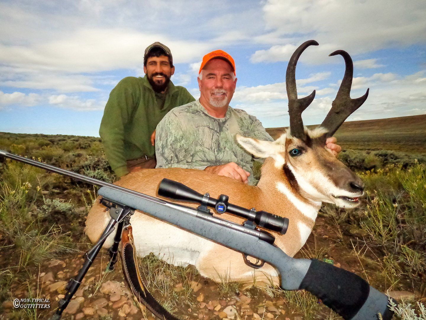 Two men pose with a pronghorn antelope in a field.
