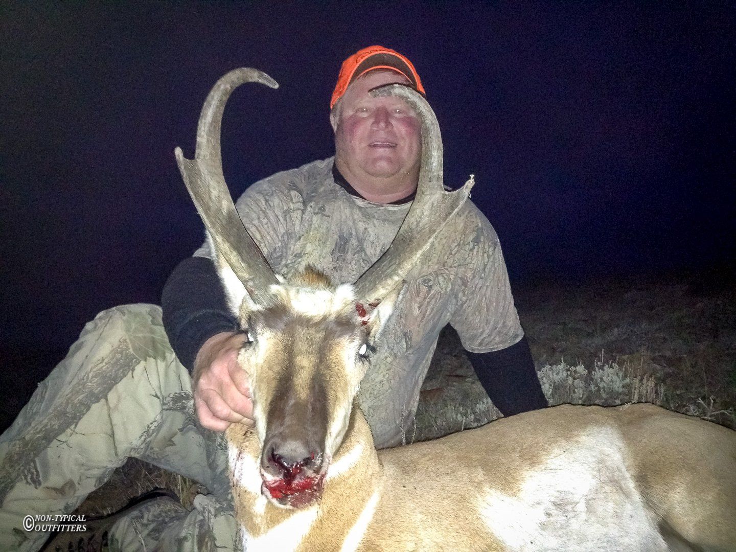 Man with orange hat holding a dead pronghorn antelope at night; bloody nose, large horns.