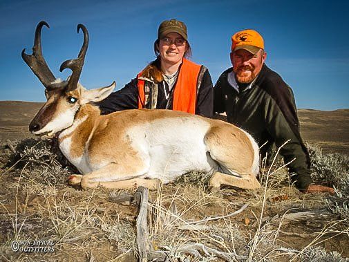 Two people pose with a pronghorn antelope on dry grassland; one wears blaze orange.