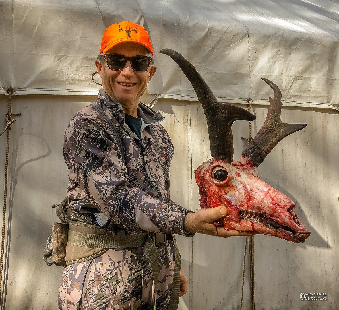 Man in camouflage showing trophy buck antelope smiling. White tent in the background.