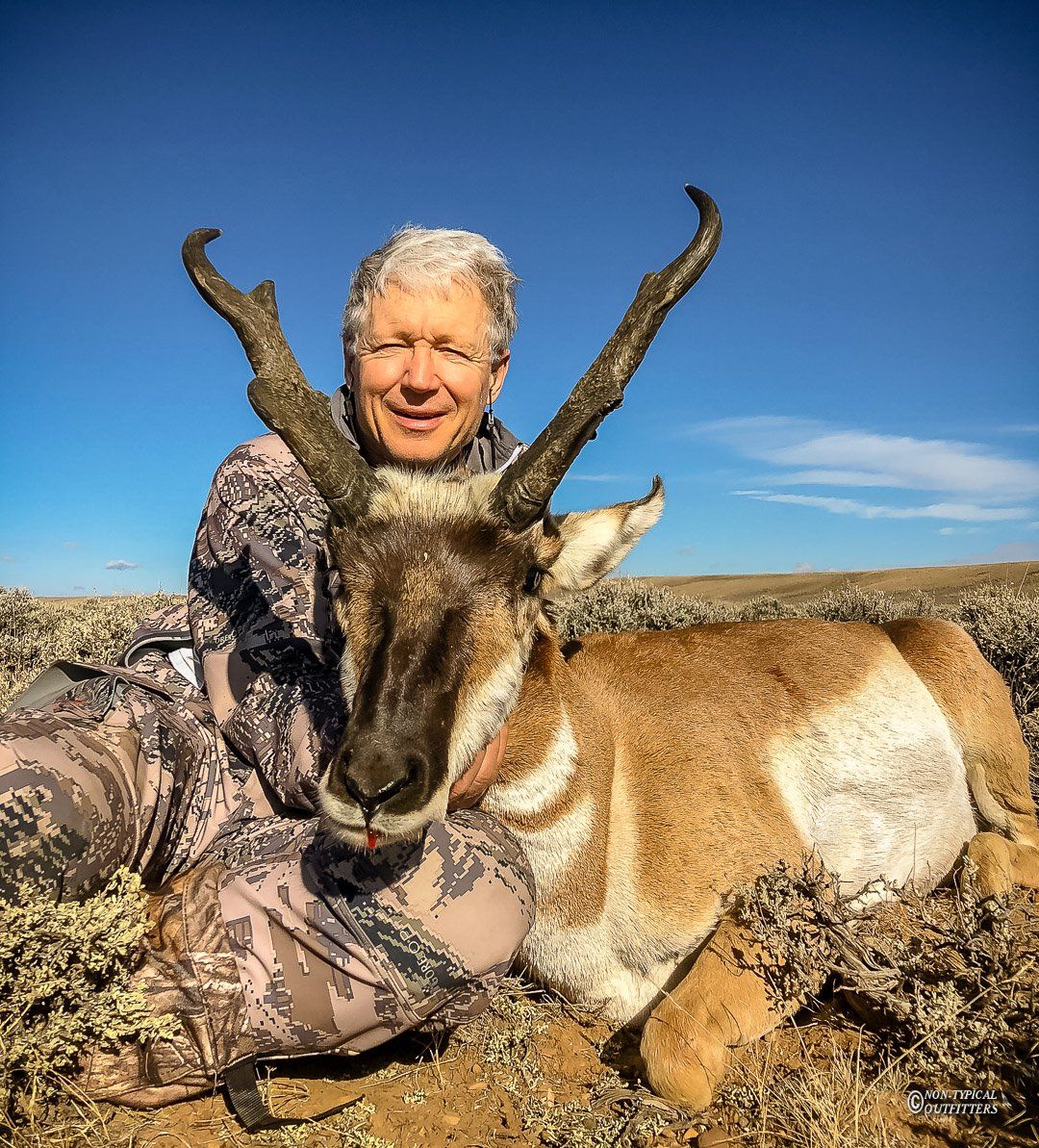 Man kneels with a pronghorn antelope in a field under a blue sky; wearing camouflage, smiles.