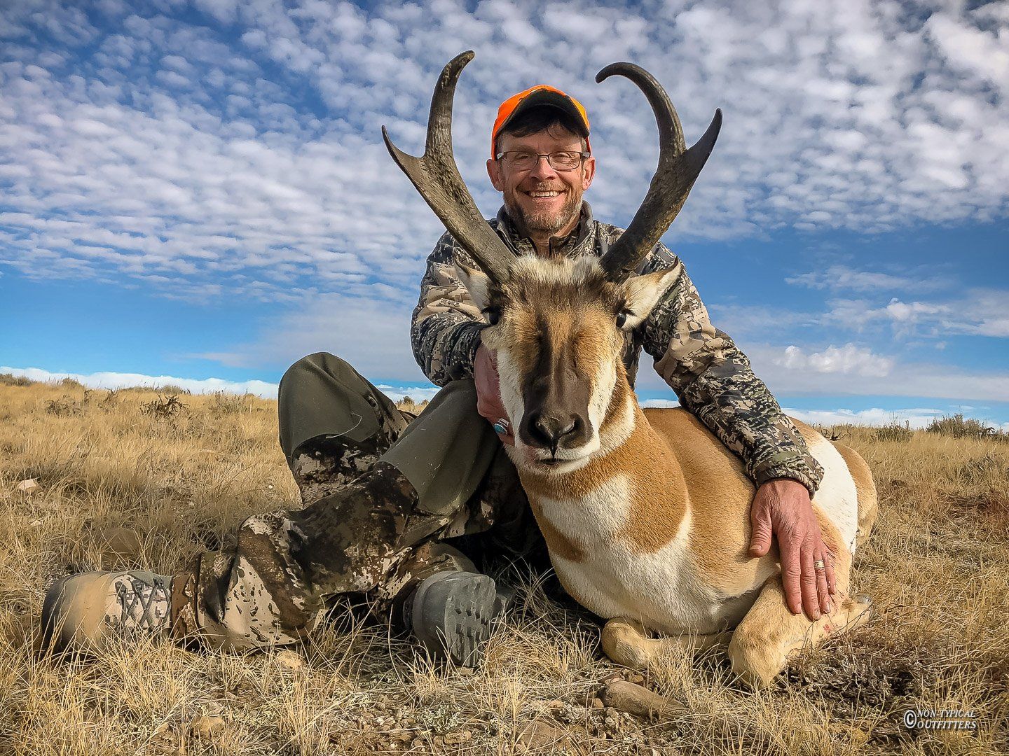 Hunter kneels with a pronghorn antelope in a field. Man wears camouflage and an orange hat, smiling.
