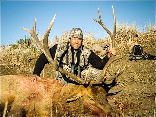 Hunter in camouflage with a large elk, outdoors. He’s smiling, holding its antlers. Blood on the elk’s chest.