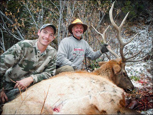 Two men pose with a large elk in a snowy forest. One man wears camo, the other a hat.
