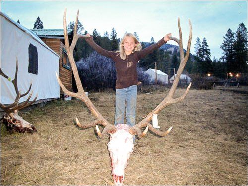 Girl standing with arms raised, atop large elk skull with antlers, outdoors.