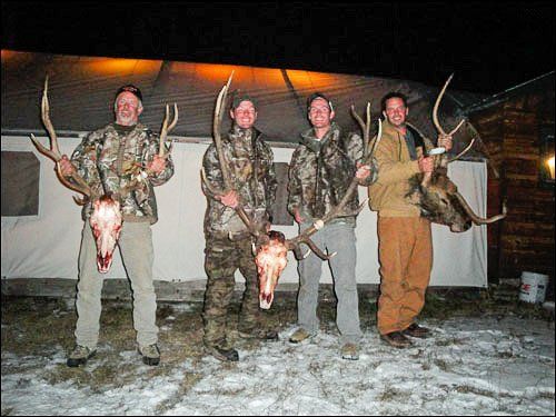 Four hunters posing with deer antlers and skulls at night. They stand in front of a white tent on snow.