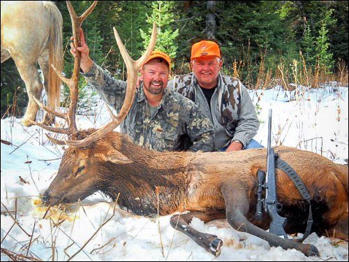 Two men in hunting gear pose with a dead elk in snow; one holds a large antler.