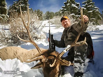 Hunter kneels in snow next to a large elk with antlers, holding the antlers up. Rifle nearby, snowy trees.