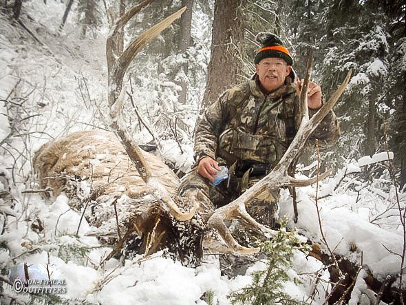 Man in camouflage sits next to a large elk in a snowy forest; man holds up a finger.