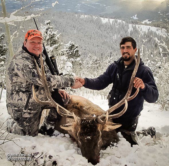 Two men shaking hands over a dead elk in snowy mountains. One man holds a rifle; the other holds antlers.