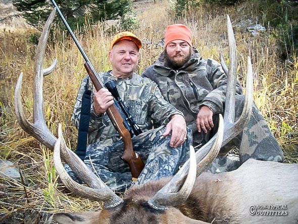 Two men in camouflage pose with a large elk and rifle. Orange hats, outdoor setting.