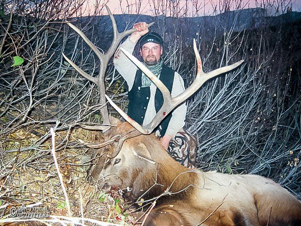 Hunter with a large elk, outdoors. Hunter kneels, holding up antlers; elk lies on the ground in a forest.