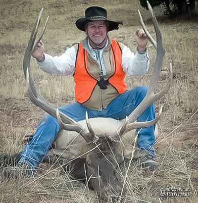 Man in hunting gear sits on a fallen elk, holding its antlers in an outdoor setting.