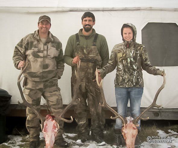 Three people in camouflage gear pose with deer skulls and antlers. Outdoor setting, snow on the ground.