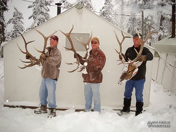 Three men in hunting gear pose with large antlers in front of a snow-covered tent.