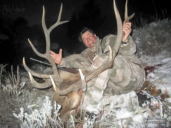 Hunter in camouflage kneels by elk with large antlers, celebrating successful hunt in snowy landscape.