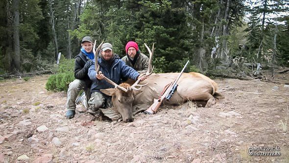 Three people pose with a dead elk in a forest clearing. The men hold rifles, smiling with triumph.