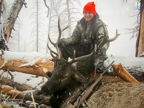 Hunter in camouflage and orange hat, kneeling with a harvested elk in a snowy forest.