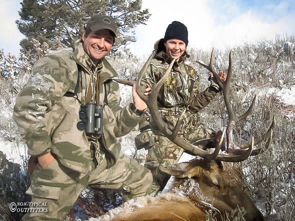 Two people in camouflage with a harvested elk in snowy mountains.