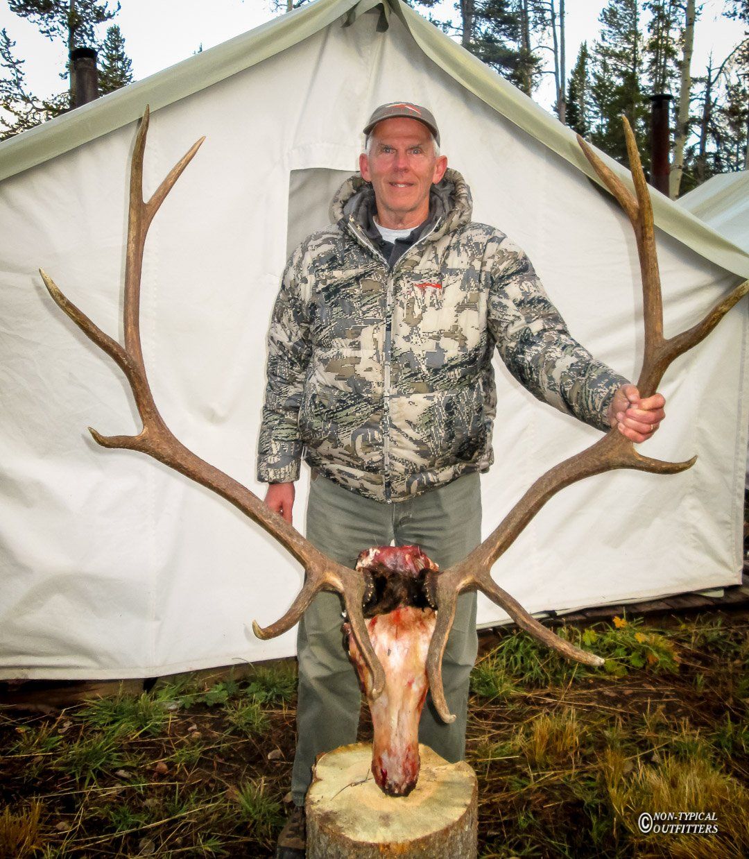 A man in a camo jacket is holding a deer skull in front of a tent