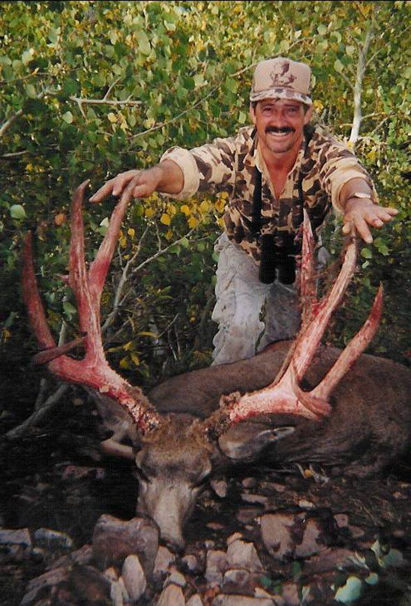 Hunter smiling, holding large antlers of a fallen deer. Outdoor setting, camouflage clothing.