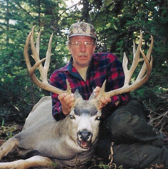 Man in plaid shirt holding large deer antlers, deer lying on ground in forest.