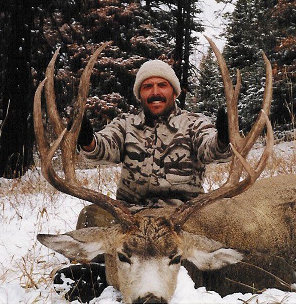 Man in camo with large deer antlers in snow. He is smiling and has his arms up.