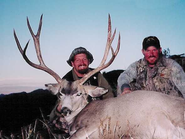 Two men pose with a large buck with impressive antlers outdoors. They are smiling.