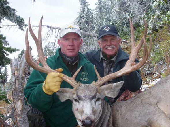Two men pose with a large buck deer, antlers prominent. Green jackets, outdoor setting.