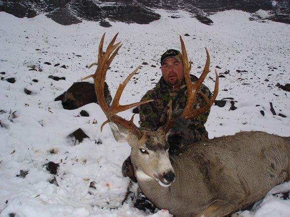Hunter in camouflage kneels in snow next to a large deer with large antlers. Mountains in the background.