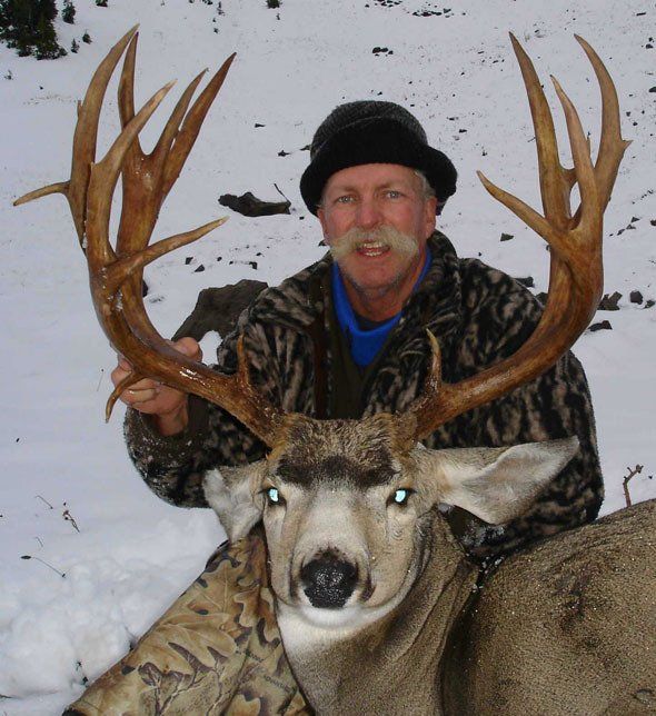 Man holding large deer antlers, snow-covered landscape. Man wearing camo jacket and hat, smiling with deer.
