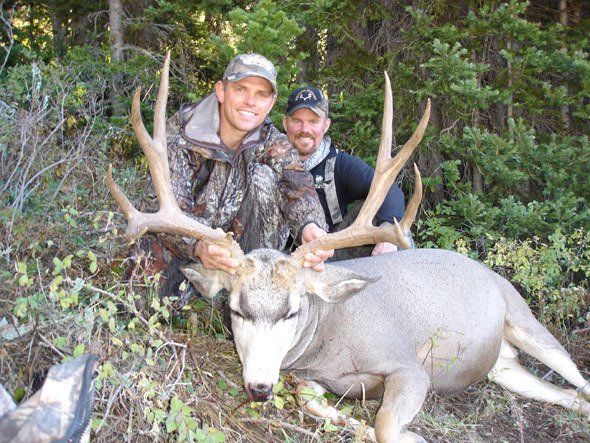 Two men pose with a large buck in a forest setting; one man kneels, holding the antlers.